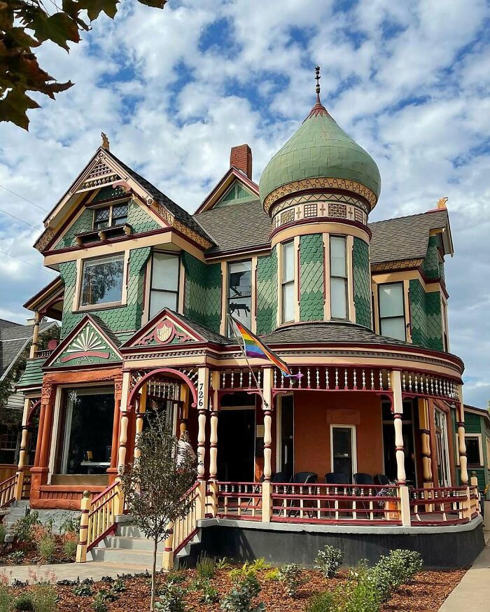 Victorian-style old home with green and red exterior, ornate detailing, and a rounded turret under a partly cloudy sky.