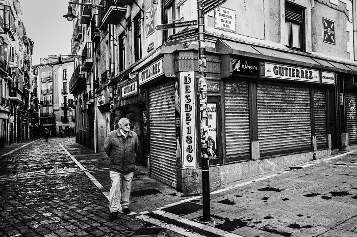 Black-and-white street photo of an elderly man walking on a cobblestone road near closed storefronts.
