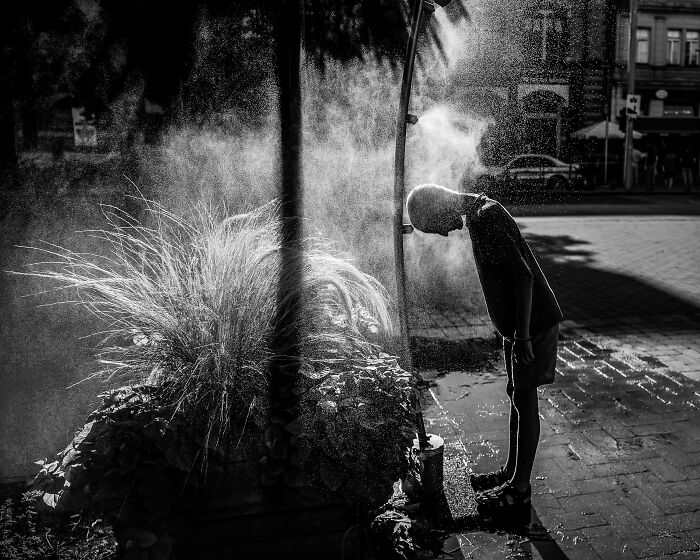Black-and-white photo of a child standing under a misting fountain in an urban setting, award-winning exposure.