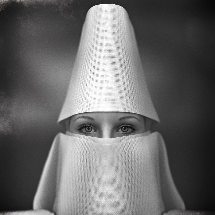 Black-and-white portrait of a person with a tall textured hat and fabric covering the face, eyes gazing directly at camera.