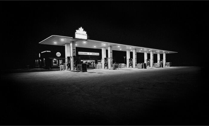 Black-and-white photo of a gas station at night with bright lights highlighting the building and pumps.