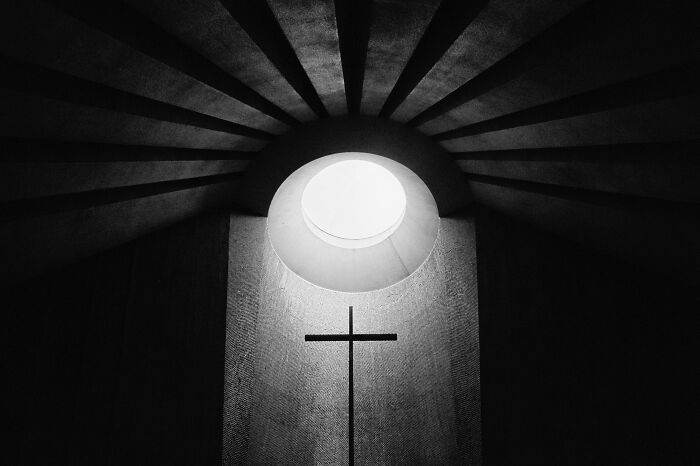 Black-and-white photo of a cross illuminated by circular light from above with geometric shadows radiating outward.