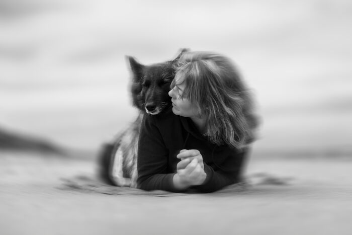 Woman lying on sand embracing her dog in a heartwarming pet photo from the 2025 International Pet Photography Awards.