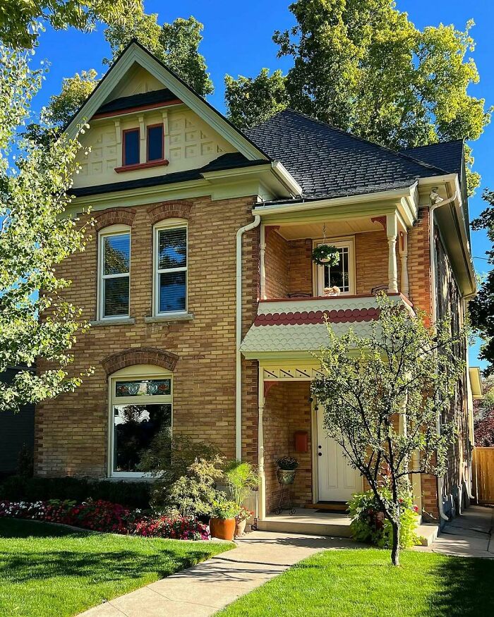 Charming old home with brick exterior, ornate windows, and a cozy front porch surrounded by greenery and flowers.
