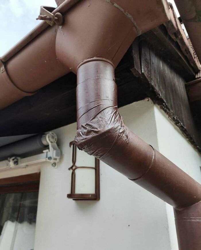Poorly repaired brown rain gutter corner wrapped with tape on the side of a white house showing flawed construction design.