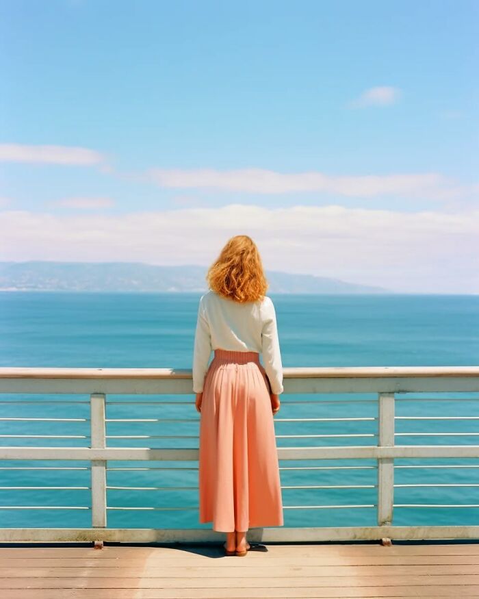 Woman in a flowing skirt standing on a deck overlooking calm ocean waves in a dreamy minimalist photo scene.
