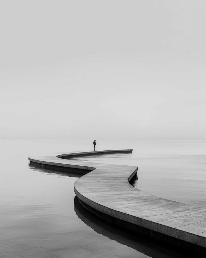 Minimalist photo of a curved walkway extending over calm water with a solitary figure, evoking a peaceful escape.