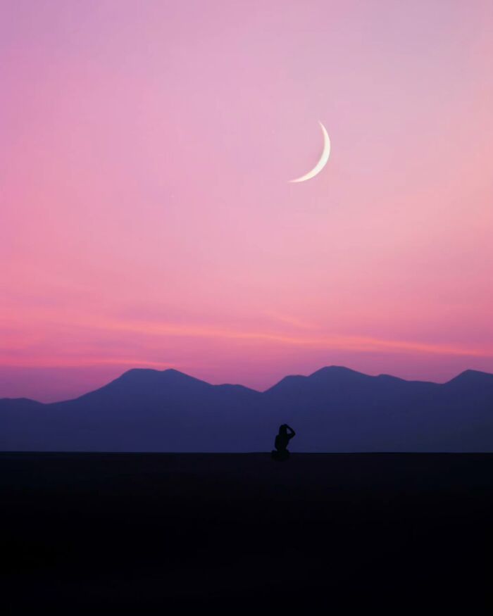 Silhouette of person sitting under a crescent moon with mountain backdrop in dreamy minimalist peaceful escape photo.