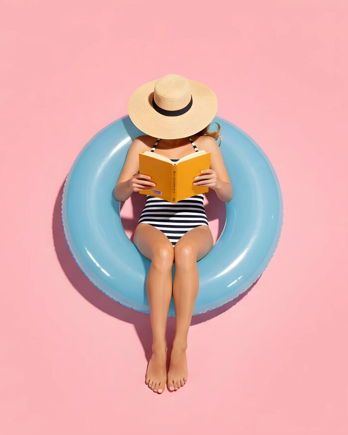 Woman in striped swimsuit and sunhat reading book while relaxing on blue float on pink background, minimalist photo.