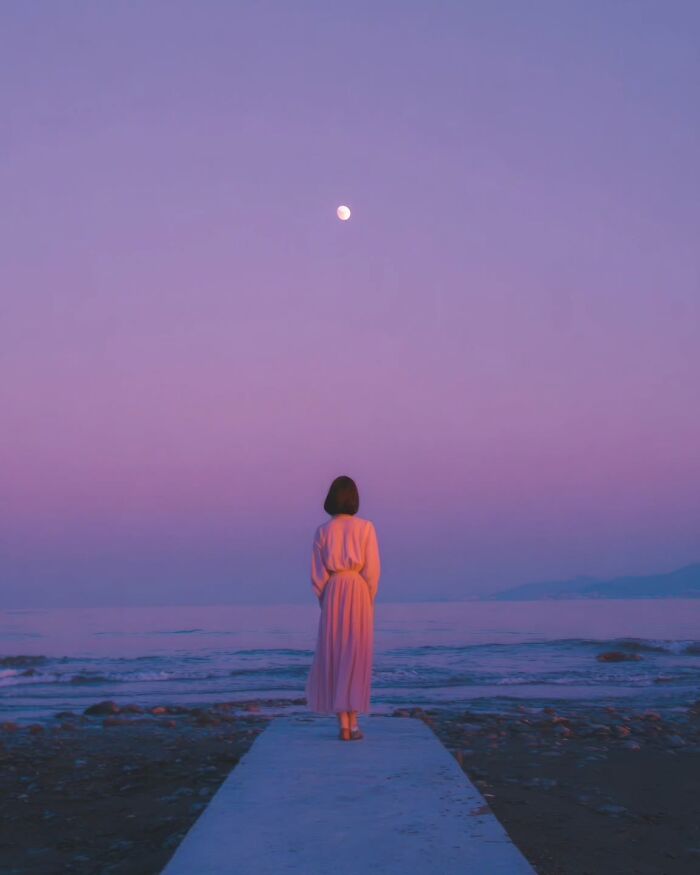 Woman in a pink dress standing by the ocean under a purple sky and moon, minimalist photo evoking peaceful escape.