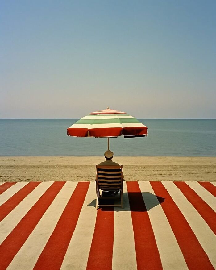 Minimalist photo of a person relaxing under a striped umbrella by the calm sea, evoking a peaceful escape.