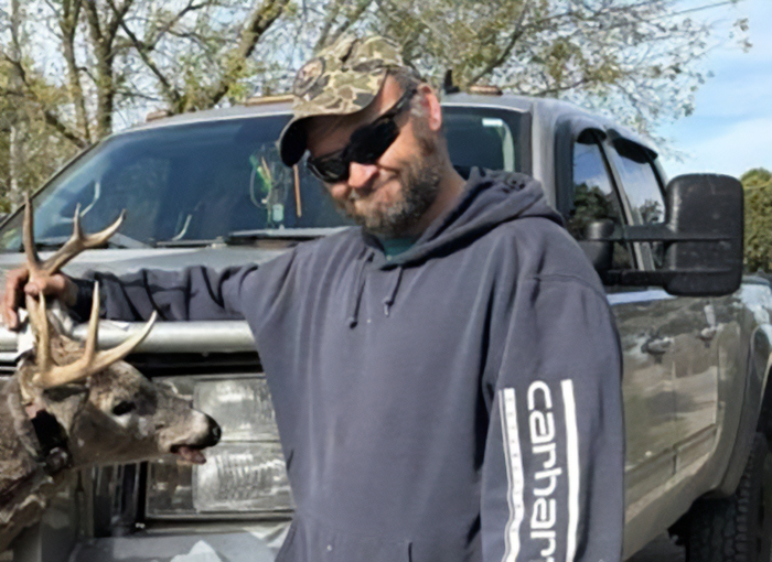 Man in sunglasses and camo cap standing by a truck with a deer, linked to Michigan church tragedy conspiracy theorists. Man in sunglasses and camo cap standing by a truck with a deer, linked to Michigan church tragedy conspiracy theorists.