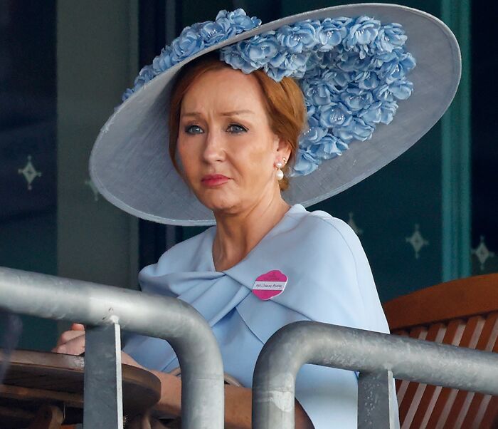 J.K. Rowling wearing a large floral hat, looking serious during an outdoor public event. J.K. Rowling wearing a large floral hat, looking serious during an outdoor public event.