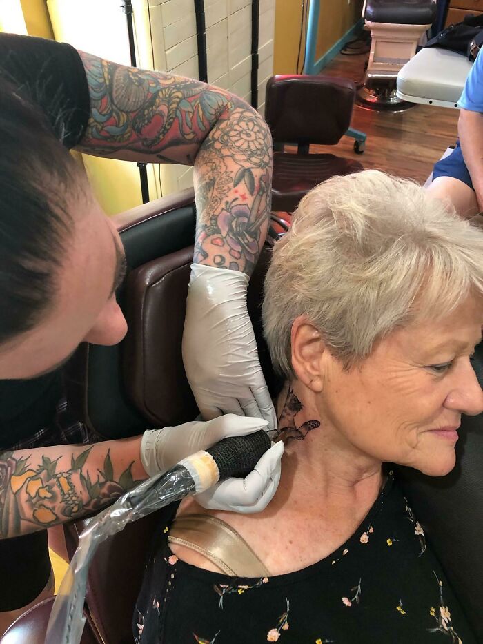 Tattoo artist with colorful sleeve ink works on an older woman’s neck tattoo in a busy studio setting.