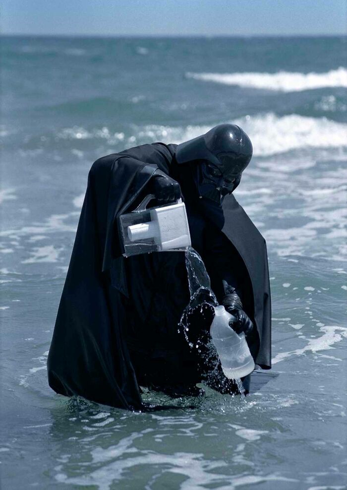 Person dressed in a dark costume pouring water from a pitcher into a glass, standing in the ocean waves, strange unexplained photo.