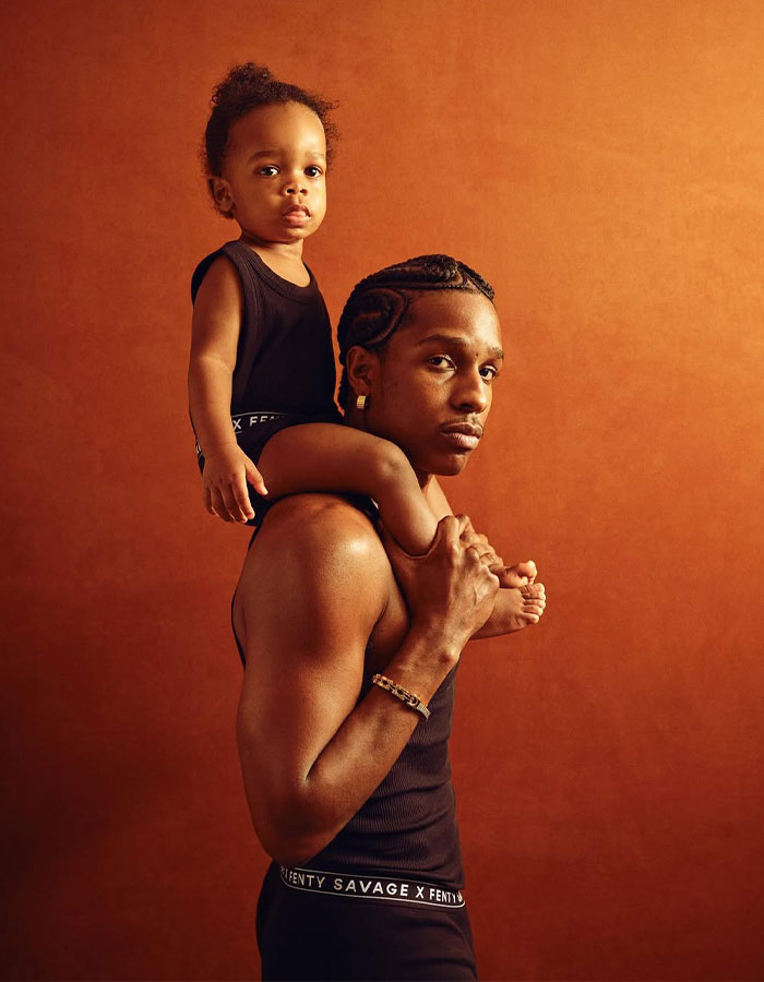 Man carrying toddler on shoulders wearing matching black Fenty Savage outfits in front of brown backdrop related to Rihanna fans urge cruel name. Man carrying toddler on shoulders wearing matching black Fenty Savage outfits in front of brown backdrop related to Rihanna fans urge cruel name.