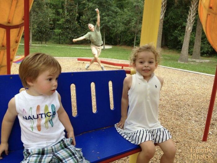 Two kids sitting on a swing at a playground while their funny parent photobombs in the background.