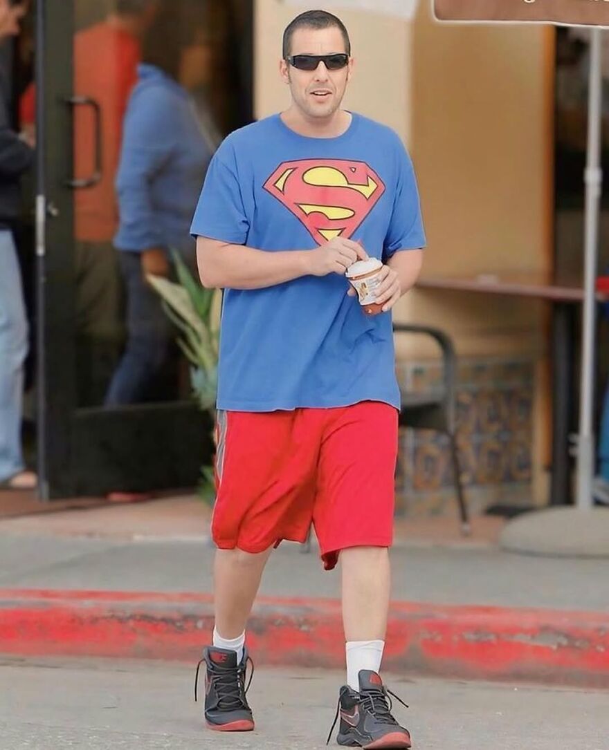 Adam Sandler wearing a casual outfit with a Superman shirt, red shorts, and sunglasses walking outdoors.