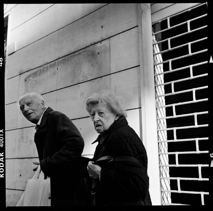 Black and white analog photo of an elderly couple walking outdoors, captured in a candid street photography style.