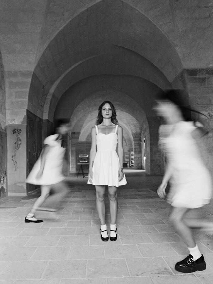 Black and white analog photo of a woman in a white dress standing still while two others move around her in an archway.