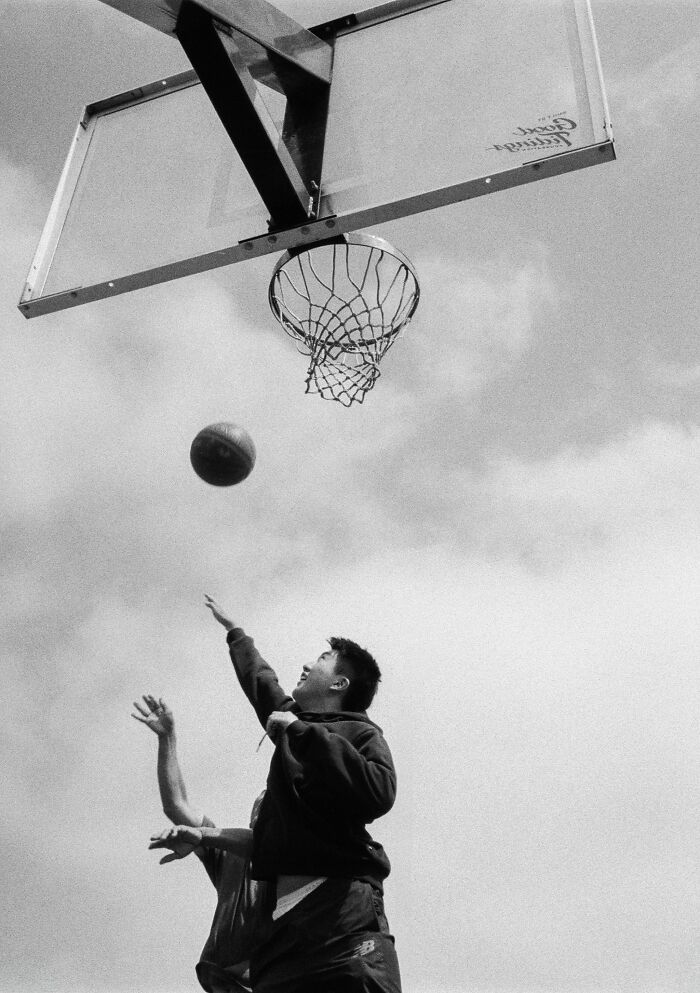 Black and white analog photo of two people playing basketball outdoors under a cloudy sky.