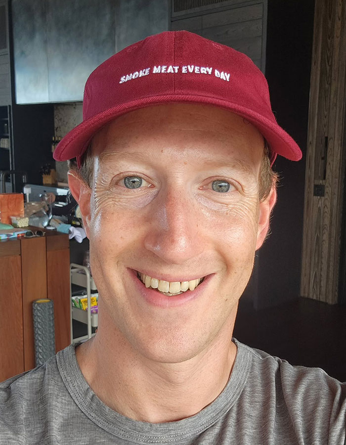 Mark Zuckerberg wearing red cap and gray shirt, smiling in a casual indoor setting related to hot mic moment. Mark Zuckerberg wearing red cap and gray shirt, smiling in a casual indoor setting related to hot mic moment.
