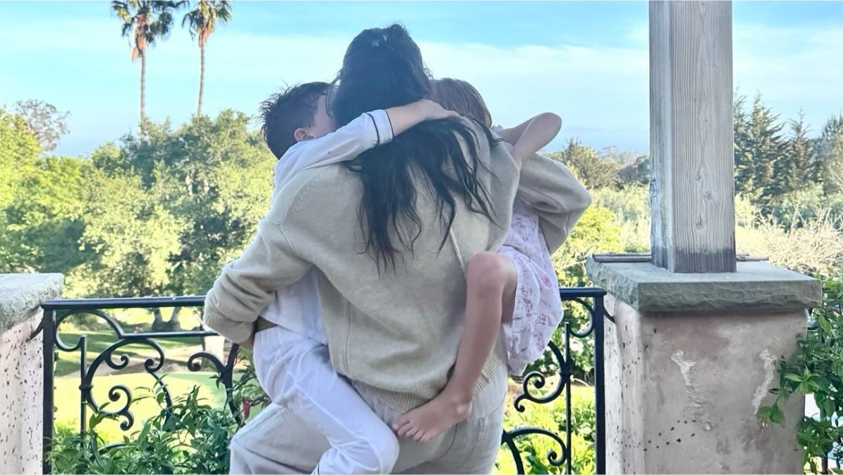 Meghan Markle hugging Prince Harry's kids outdoors on a balcony with scenic greenery in the background.
