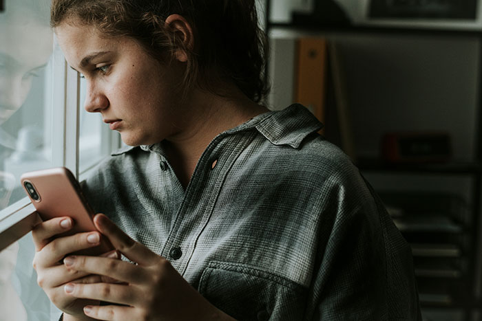 Teen girl looking troubled, holding a smartphone by a window as her life is shaken after parents' long world trip return. Teen girl looking troubled, holding a smartphone by a window as her life is shaken after parents' long world trip return.