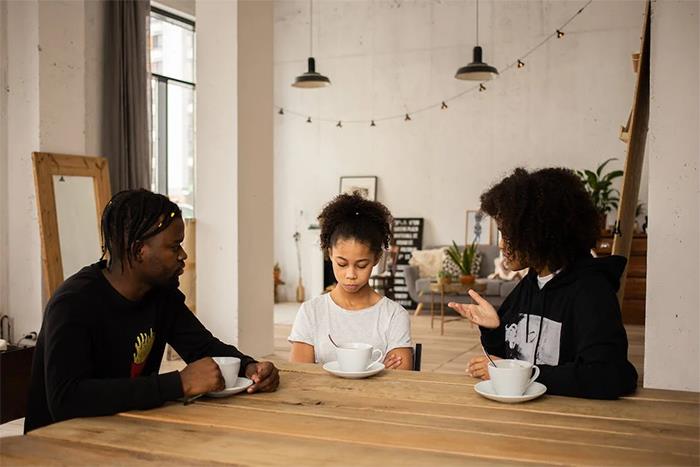 Teen girl looking upset while parents talk to her at a wooden table after returning from a long world trip. Teen girl looking upset while parents talk to her at a wooden table after returning from a long world trip.