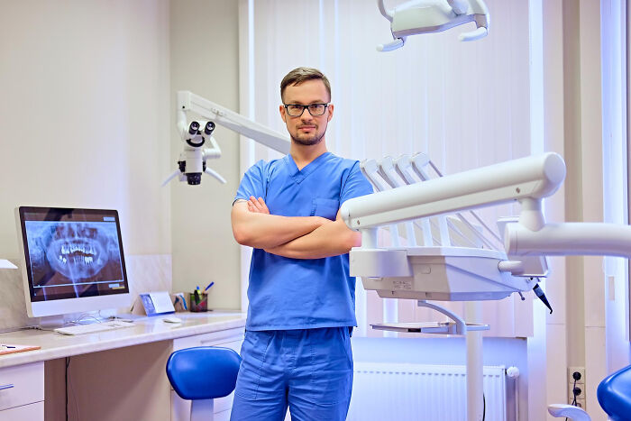 Dentist in blue scrubs standing in clinic with arms crossed near dental chair and X-ray screen showing teeth. Dentist in blue scrubs standing in clinic with arms crossed near dental chair and X-ray screen showing teeth.