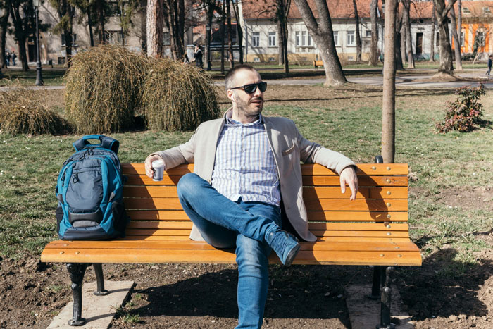 Man wearing sunglasses sitting on a park bench with backpack nearby, in a no-walking area setting. Man wearing sunglasses sitting on a park bench with backpack nearby, in a no-walking area setting.