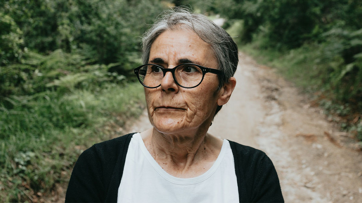 Older adult woman with glasses standing on a forest path, showing a serious expression, capturing adults who failed to grow up.