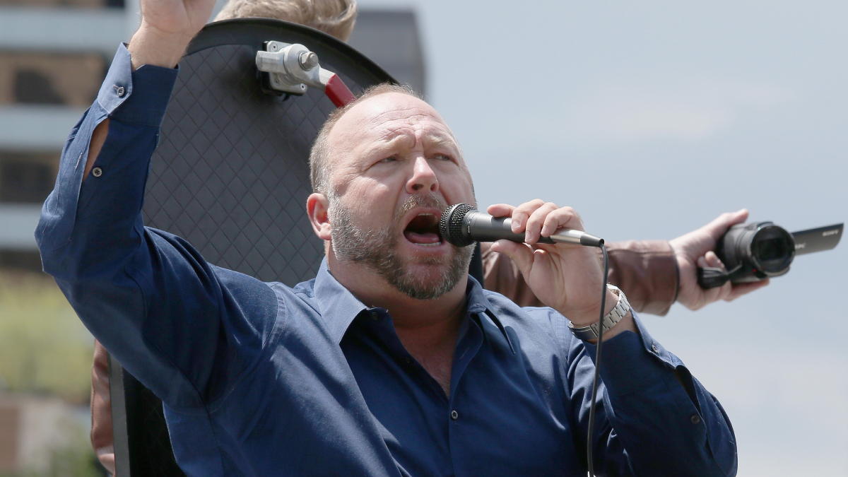 Alex Jones passionately speaking into a microphone outdoors with a crowd and camera equipment behind him.
