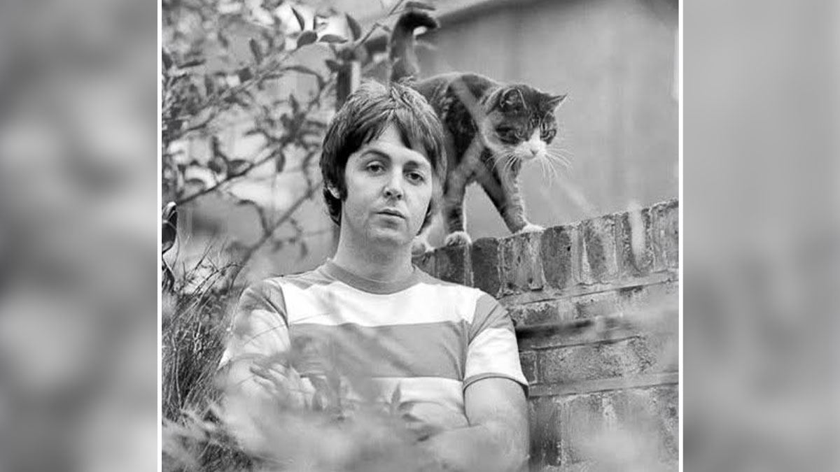 Black and white vintage cat photo featuring a man sitting near a brick wall with a cat walking behind him outdoors.