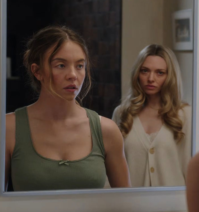 Amanda Seyfried looking serious in a mirror reflection with a woman in a white sweater behind her indoors. Amanda Seyfried looking serious in a mirror reflection with a woman in a white sweater behind her indoors.
