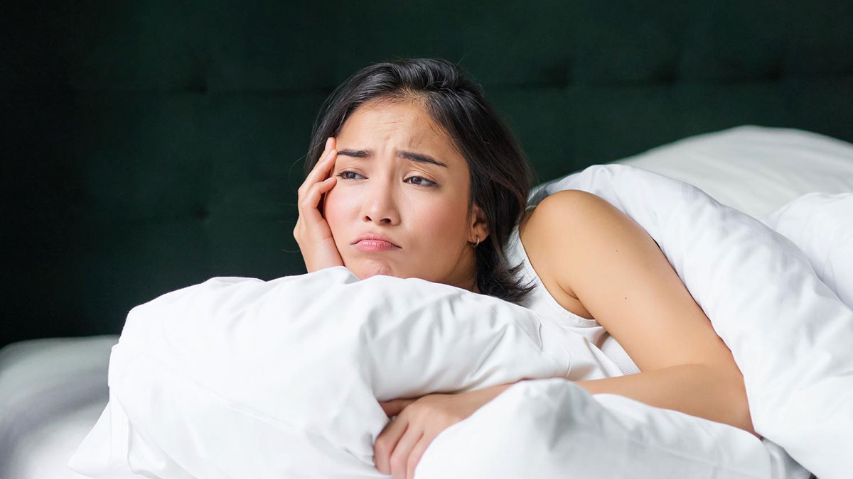Woman lying in bed looking worried and thoughtful, illustrating concerns about a throuple and an anime pillow.