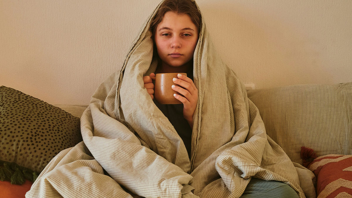Young woman wrapped in a blanket holding a mug, looking unwell and uncertain about attending wedding with positive Covid test