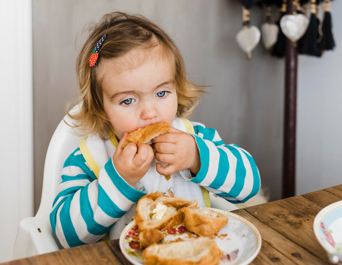 Toddler girl eating bread at a wooden table, highlighting concerns about eating disorder comments from ex and dad's reaction. Toddler girl eating bread at a wooden table, highlighting concerns about eating disorder comments from ex and dad's reaction.