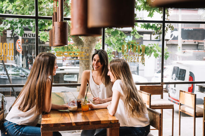 Three women sitting at a wooden table in a cafe, chatting and enjoying drinks in a bright, leafy setting. Three women sitting at a wooden table in a cafe, chatting and enjoying drinks in a bright, leafy setting.