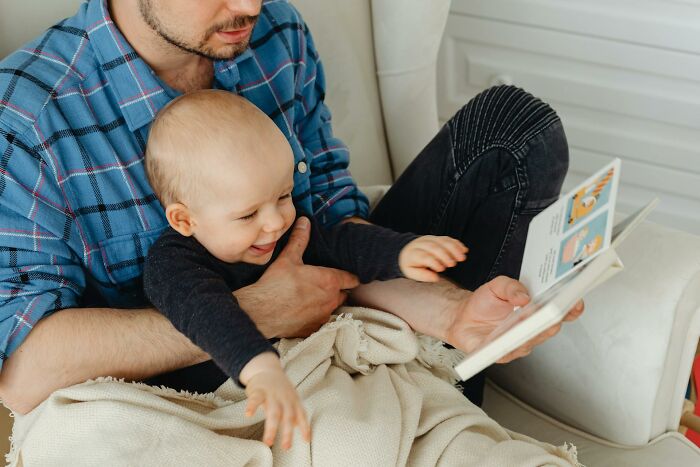 Dad taking care of son for the first time, reading a book while holding baby wrapped in a beige blanket at home. Dad taking care of son for the first time, reading a book while holding baby wrapped in a beige blanket at home.