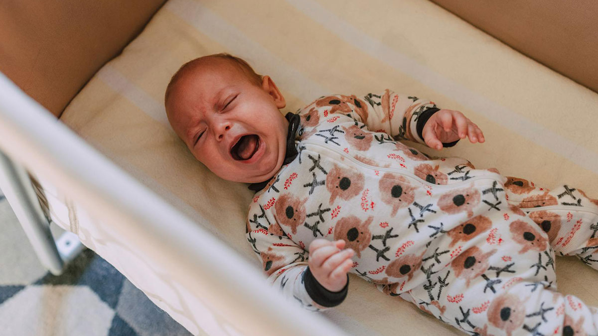Crying baby boy lying in crib wearing bear-patterned pajamas, showing dad's first-time childcare challenges.