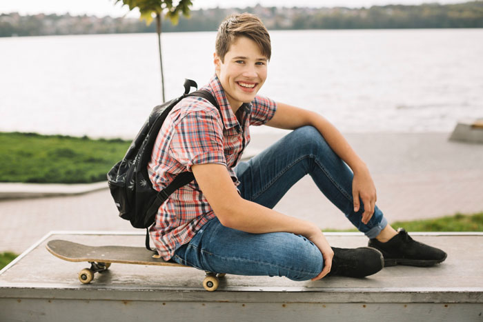 Teen boy with backpack sitting on skateboard by the water, representing woman’s teen bro who is a nightmare to deal with. Teen boy with backpack sitting on skateboard by the water, representing woman’s teen bro who is a nightmare to deal with.
