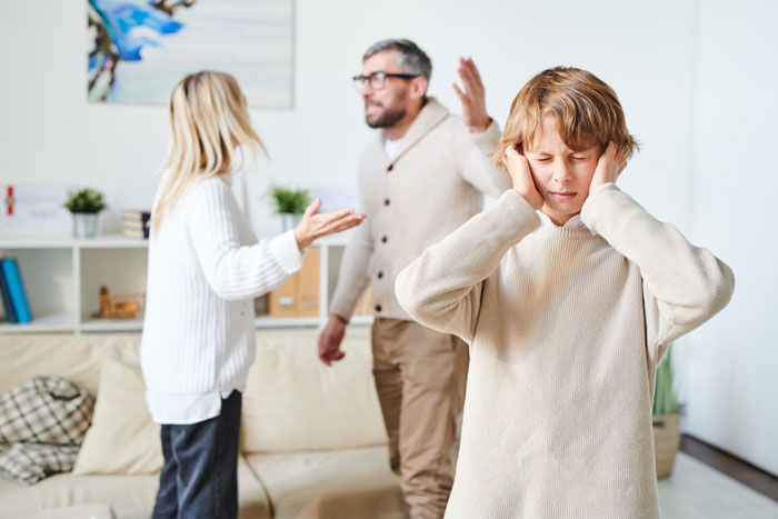Teen boy covering ears while parents argue in the background about babysitting and family conflict tensions. Teen boy covering ears while parents argue in the background about babysitting and family conflict tensions.