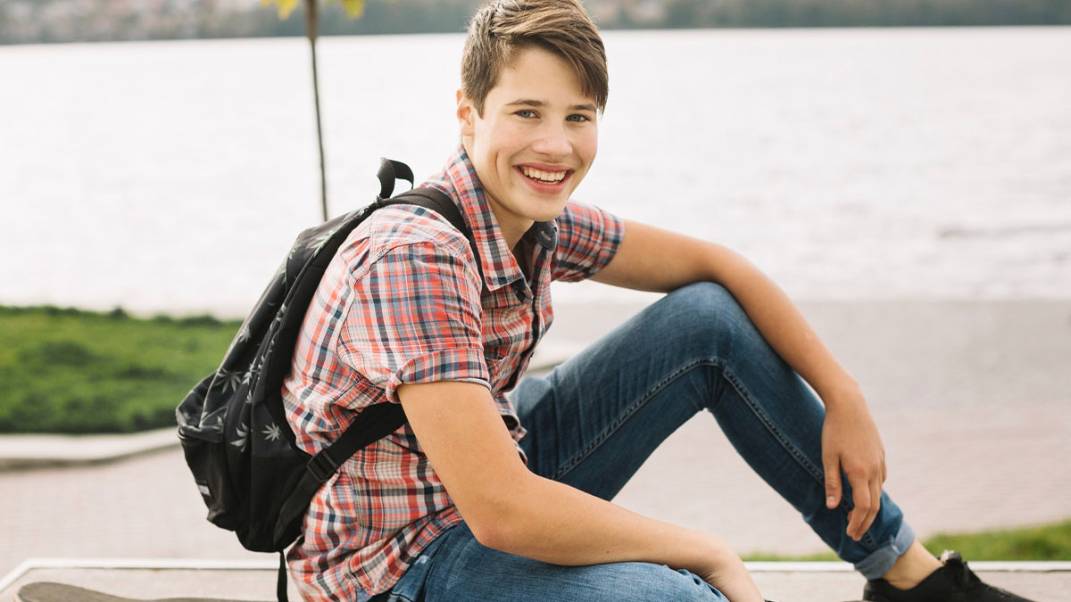 Teen boy wearing a backpack and plaid shirt sitting by the water, representing a challenging teen brother situation.