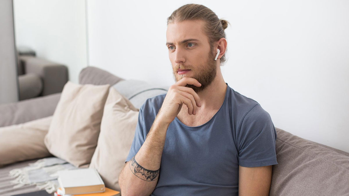 Thoughtful man with beard and earbuds sitting on couch reflecting on birth family tracking him down after abandonment.