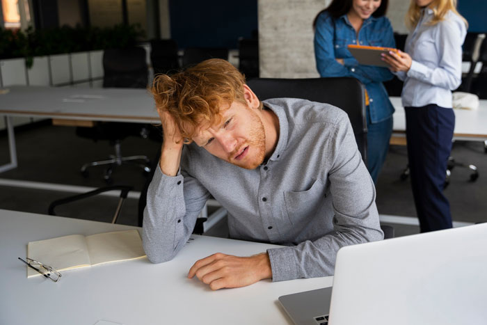 Frustrated man at office desk with laptop looking stressed while two women in background discuss boss invite vacation with her. Frustrated man at office desk with laptop looking stressed while two women in background discuss boss invite vacation with her.
