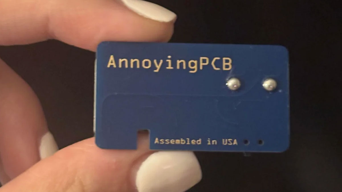 Close-up of a small blue hidden device labeled AnnoyingPCB held between fingers, related to prank and hidden device discovery.