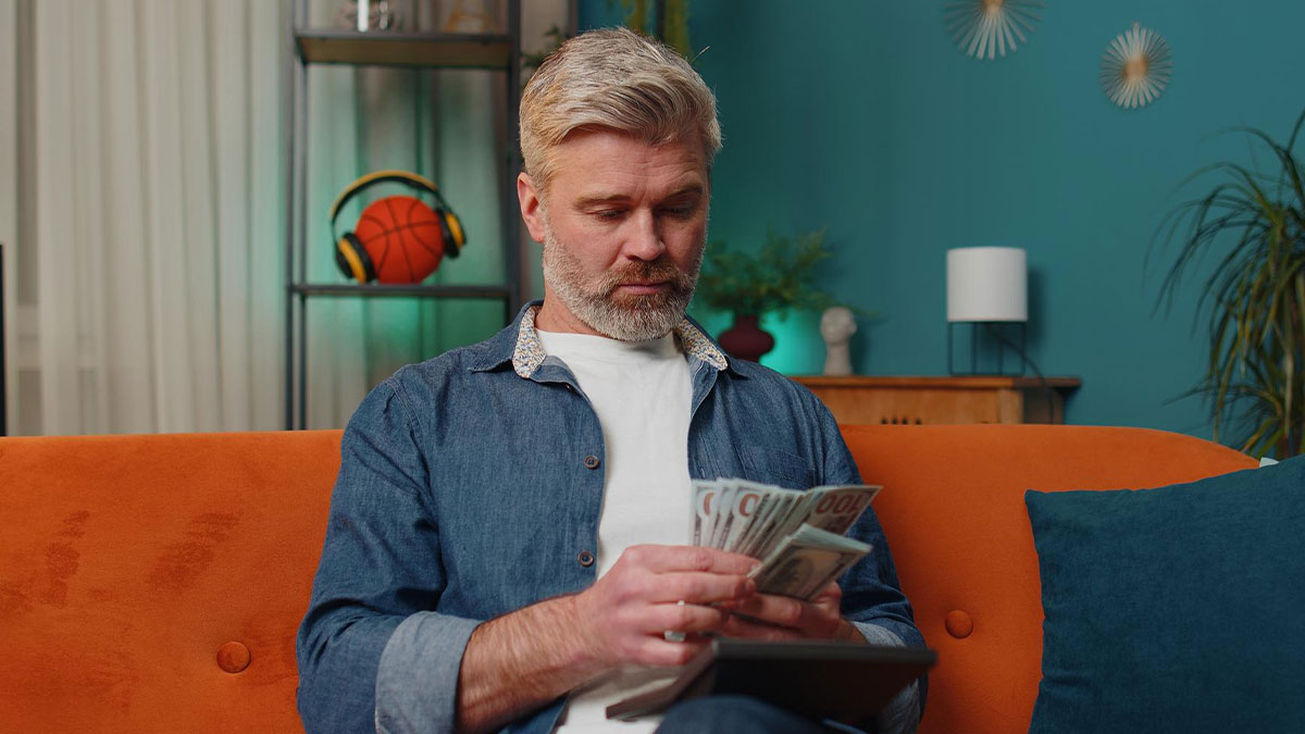 Man counting money on an orange couch, depicting greed related to sonu2019s survivor benefits in a home setting.
