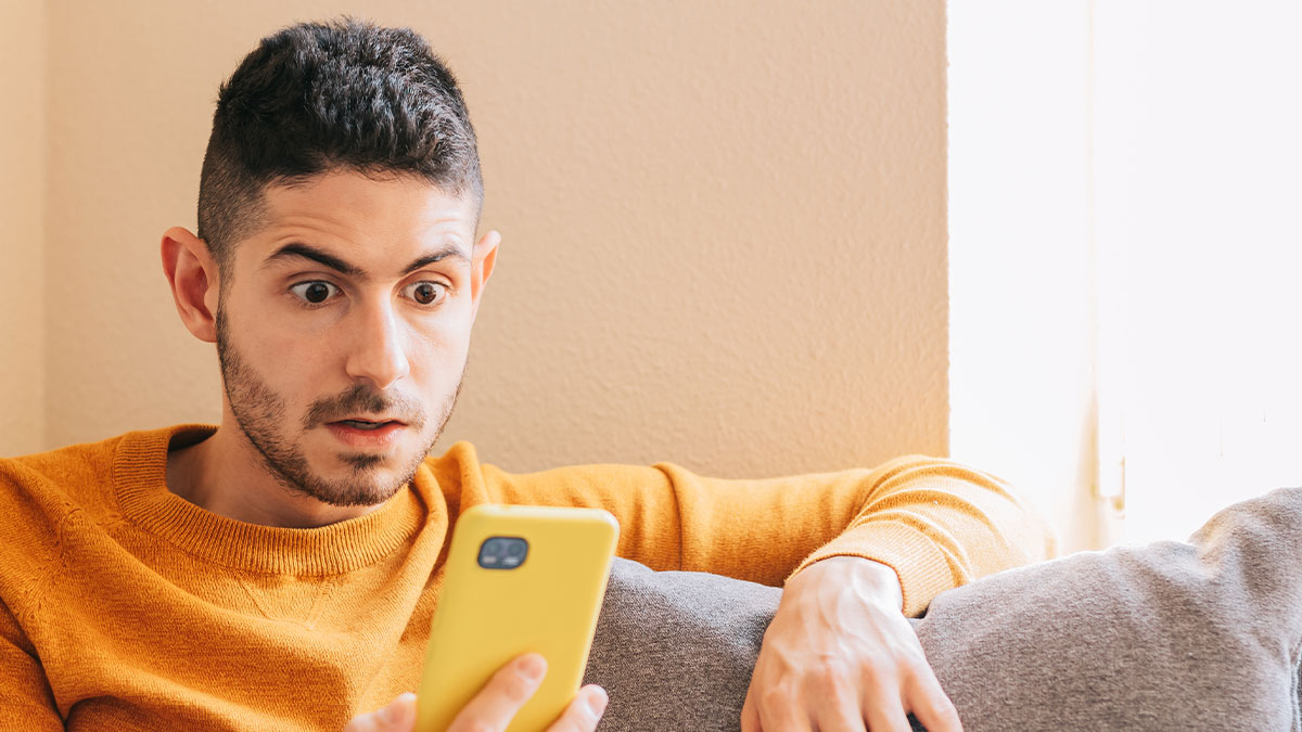 Man looking shocked at phone screen while sitting on couch, illustrating man goes through girlfriendu2019s phone scenario.
