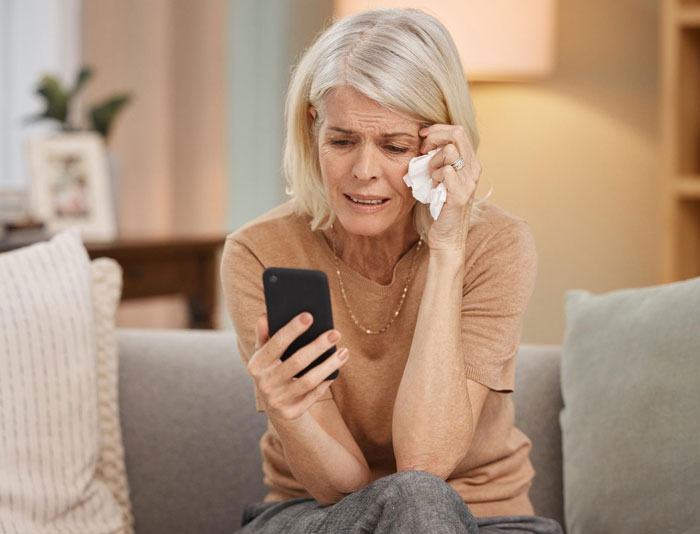 Older woman sitting on a sofa crying while looking at her phone, reflecting emotions related to breaking up over photograph. Older woman sitting on a sofa crying while looking at her phone, reflecting emotions related to breaking up over photograph.